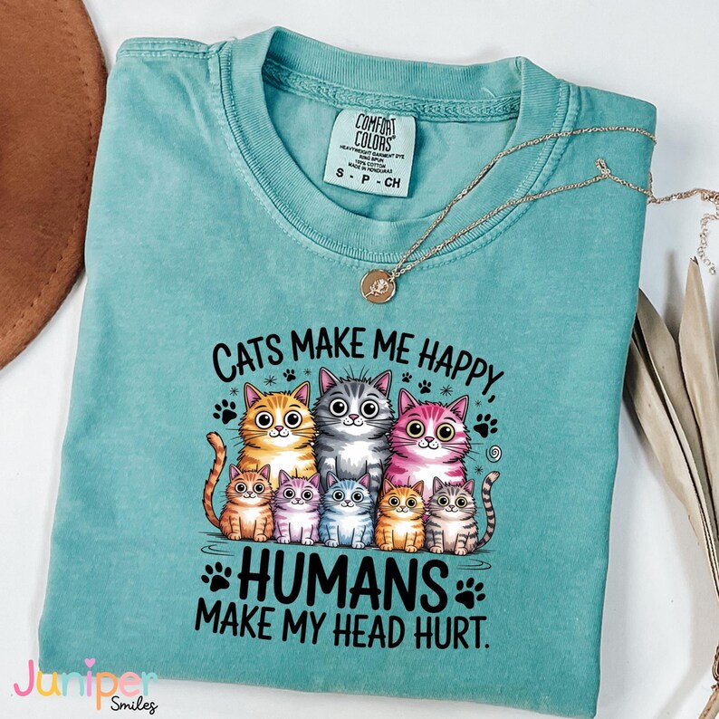 Puede incluir: Una camiseta turquesa Comfort Colors con el texto "Cats Make Me Happy, Humans Make My Head Hurt". La camiseta presenta un gr&aacute;fico de varios gatos de dibujos animados. Un collar dorado con un colgante est&aacute; drapeado sobre la camiseta.