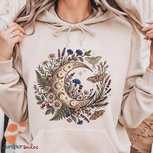 May include: Beige hoodie featuring a crescent moon design with floral and butterfly accents. The moon is surrounded by daisies, lavender, and other flowers. The text "Juniper smiles" is at the bottom left.