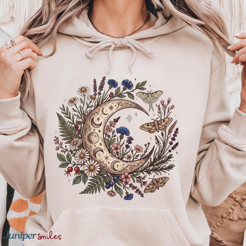 May include: Beige hoodie featuring a crescent moon design with floral and butterfly accents. The moon is surrounded by daisies, lavender, and other flowers. The text "Juniper smiles" is at the bottom left.