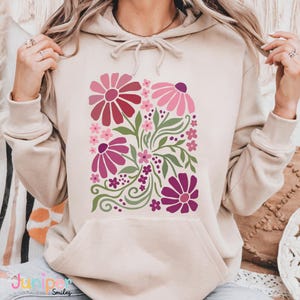 May include: A beige hoodie featuring a floral design in shades of pink and purple. The design includes stylized flowers, leaves, and swirls. The hoodie has a kangaroo pocket and a drawstring hood. The text "Juniper Smiles" is visible at the bottom.