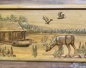 May include: A wooden carved wall art depicting a cabin in the woods with a deer drinking from a pond. Two ducks fly overhead. The scene is carved in a light brown wood with green accents.