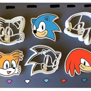 May include: Six white plastic cookie cutters shaped like the heads of characters from the Sonic the Hedgehog video game series. The characters are Sonic, Tails, Knuckles, Shadow, and two versions of  Sonic.