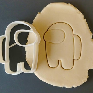 May include: White plastic cookie cutter shaped like an Among Us character. The cutter is next to a piece of dough that has been cut out in the same shape.
