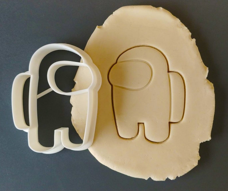 May include: White plastic cookie cutter shaped like an Among Us character. The cutter is next to a piece of dough that has been cut out in the same shape.