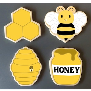May include: Four yellow and white cookie cutters shaped like a honeycomb, a bee, a honey pot, and a beehive. The honey pot cookie cutter has the word "honey" written on it.