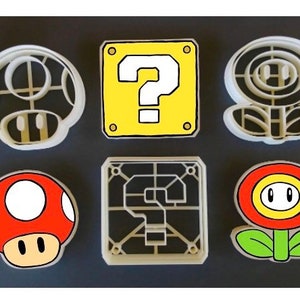 May include: Set of six white plastic cookie cutters shaped like iconic Super Mario Bros. characters and items. The cutters include a mushroom, a question block, a flower, a question block with a grid pattern, a mushroom with red and white accents, and a flower with red, yellow, and green accents.