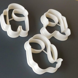 May include: Three white plastic cookie cutters shaped like the characters from the video game Among Us. The cutters are on a gray surface.