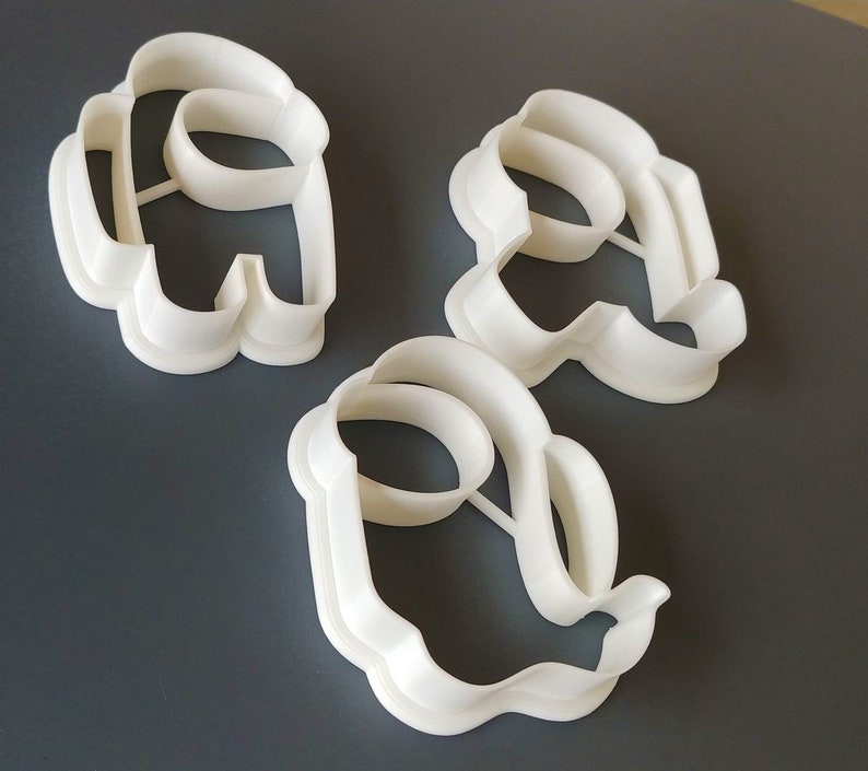 May include: Three white plastic cookie cutters shaped like the characters from the video game Among Us. The cutters are on a gray surface.