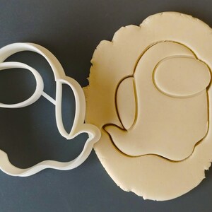 May include: A white plastic cookie cutter shaped like an Among Us character, with a round head and a single eye, next to a cookie dough cutout of the same shape.