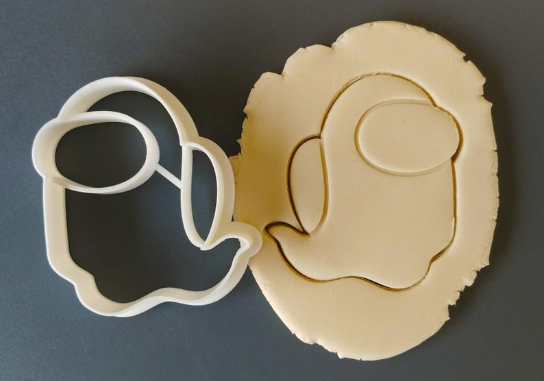 May include: A white plastic cookie cutter shaped like an Among Us character, with a round head and a single eye, next to a cookie dough cutout of the same shape.