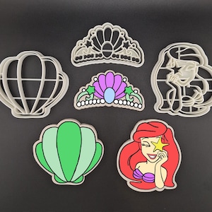May include: Set of six white plastic cookie cutters shaped like a mermaid, a seashell, a crown, and a crown with a purple jewel.