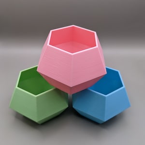 May include: Three geometric planters in pink, green, and blue. The planters are hexagonal in shape and have a smooth, matte finish.