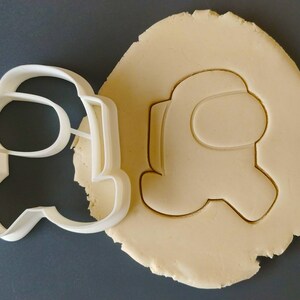 May include: A white plastic cookie cutter shaped like an Among Us character. The cutter is on top of a piece of unbaked cookie dough.