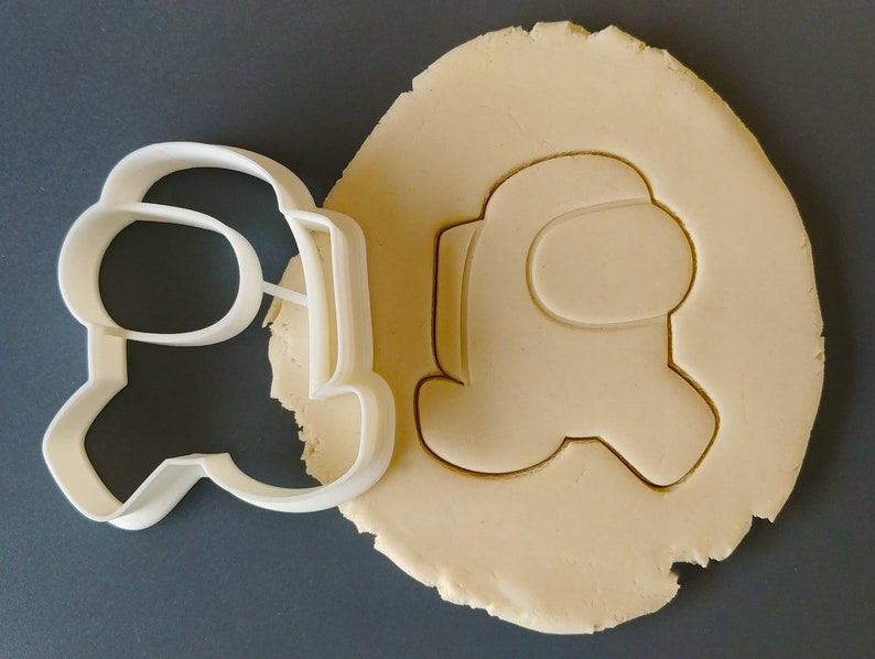 May include: A white plastic cookie cutter shaped like an Among Us character. The cutter is on top of a piece of unbaked cookie dough.