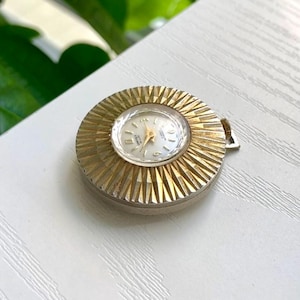 Peut inclure: Une montre de poche vintage dorée avec une lunette texturée en forme d'éclat de soleil. Le cadran de la montre est blanc avec des aiguilles et des chiffres dorés. La montre est ronde et possède une petite couronne pour le remontage.