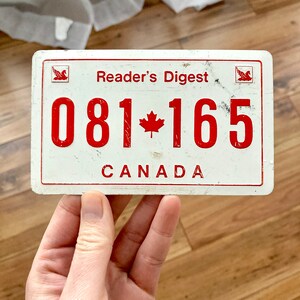 Peut inclure: Plaque en métal blanc avec du texte rouge et une feuille d'érable rouge. Le texte indique "Reader's Digest" et "CANADA" avec le numéro "081-165" entre les deux.