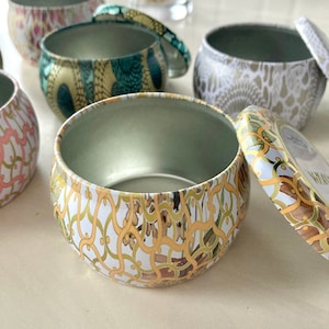 Puede incluir: Conjunto de cinco latas de velas decorativas con tapas. Las latas son redondas y presentan diferentes patrones florales y geométricos en dorado, blanco y verde.