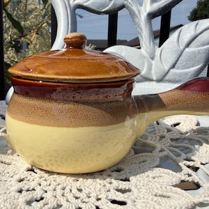Peut inclure: Un bol à soupe en céramique avec couvercle et une poignée latérale. L'objet présente un vernis tricolore, passant d'un couvercle brun foncé à une section centrale brun moyen moucheté, et une base crème unie à la finition brillante.