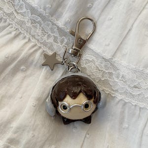 May include: A handmade keychain featuring a cartoon character with brown hair, glasses, and earmuffs. The character is attached to a silver-colored clasp and a star charm. The background is a white, textured fabric.