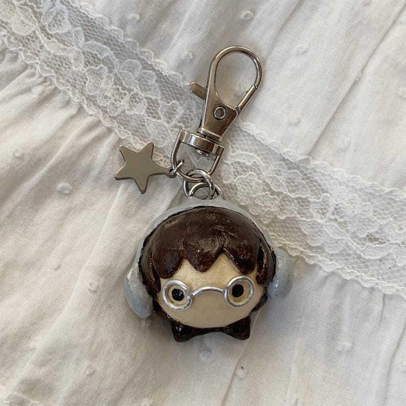 May include: A handmade keychain featuring a cartoon character with brown hair, glasses, and earmuffs. The character is attached to a silver-colored clasp and a star charm. The background is a white, textured fabric.
