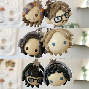 May include: A collection of handmade polymer clay keychains featuring stylized human faces. Each keychain has a unique hairstyle and color scheme, with some wearing glasses. The keychains are attached to silver-colored star-shaped charms.
