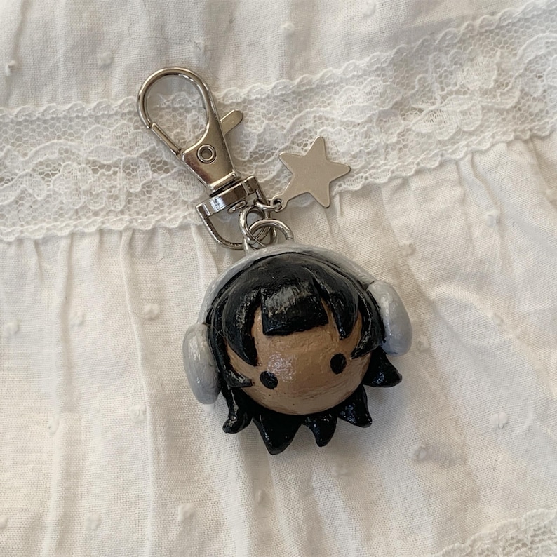 May include: A handmade keychain featuring a small, round figure with black hair, gray headphones, and a tan face. The keychain has a silver clasp and a star charm. The figure is likely made of clay or a similar material.