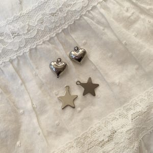 May include: Two silver heart charms and two silver star charms on a white, textured fabric. The charms are small and shiny, with a loop at the top for attaching to jewelry. The fabric has a delicate lace trim.