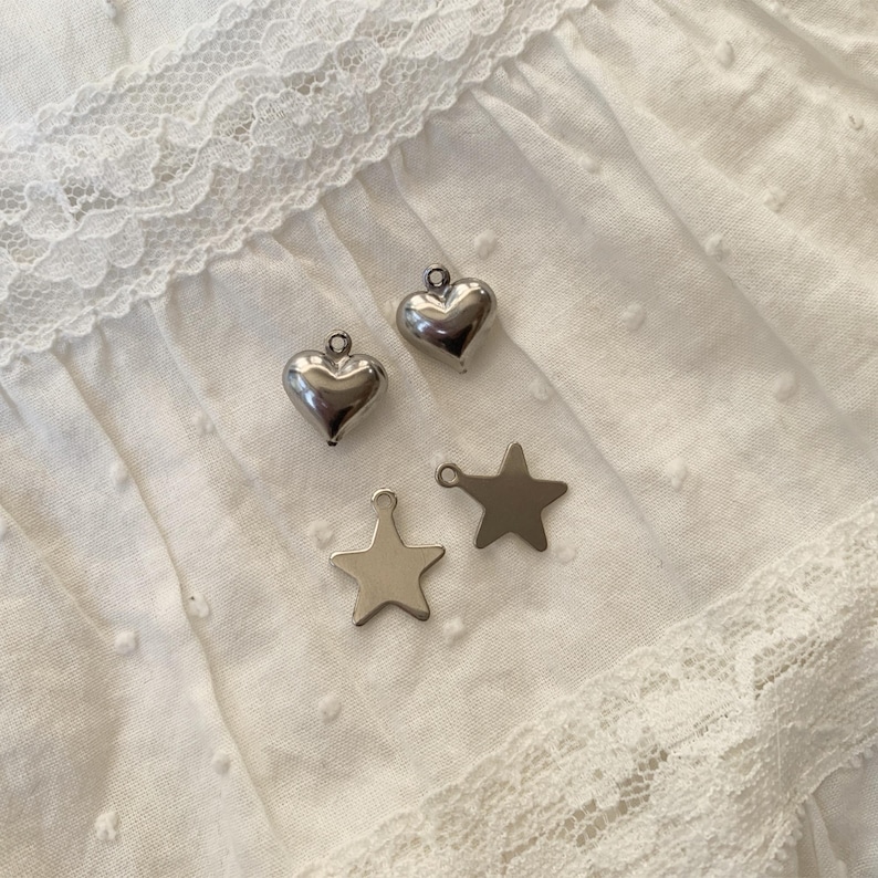May include: Two silver heart charms and two silver star charms on a white, textured fabric. The charms are small and shiny, with a loop at the top for attaching to jewelry. The fabric has a delicate lace trim.