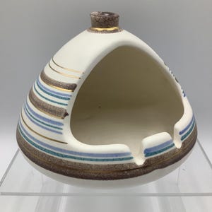 May include: Cream-colored ceramic ashtray with a unique triangular shape. The ashtray features horizontal stripes in brown, teal, and blue, accented with gold lines. The top has a small, textured brown cap. A stylish vintage piece.