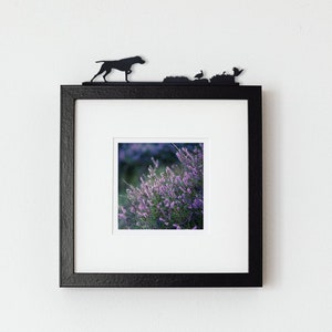 May include: Black framed picture with a white mount and a photo of purple flowers. The frame has a silhouette of a dog, birds, and bushes on the top.