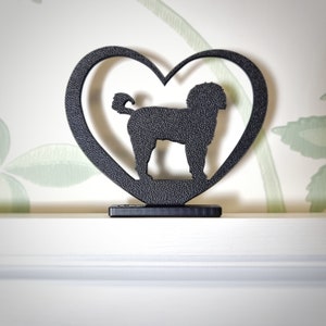 Op de afbeelding: Zwart metalen honden silhouet in een hartvormige lijst. De hond heeft een krullende staart en staat op een rechthoekige basis. De achtergrond is een witte muur met een bloemenpatroon.