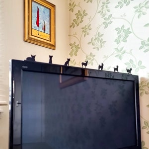 Könnte beinhalten: Ein schwarzer Flachbildfernseher mit silbernem Rahmen steht an einer Wand mit einer floralen Tapete. Auf dem Fernseher sind Silhouetten von Hunden platziert.