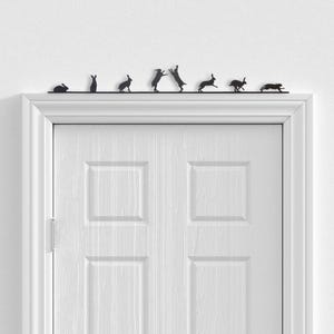 May include: A black metal door topper featuring a series of seven rabbit silhouettes in various poses, including sitting, standing, and running, positioned horizontally along a thin straight bar above a white paneled door frame.