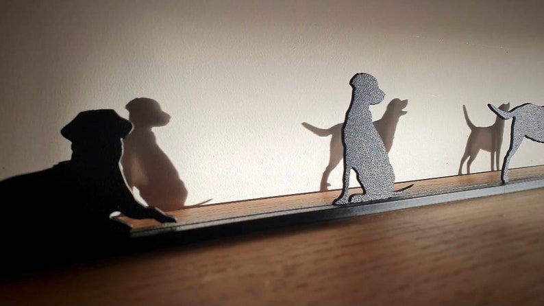 May include: A metal silhouette of four dogs standing on a black metal bar. The dogs are all facing the same direction, and their shadows are cast on the wall behind them.