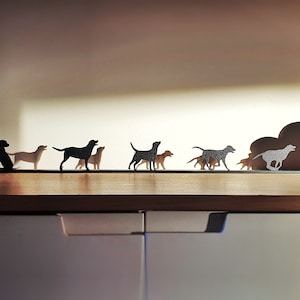 May include: A row of black metal dog silhouettes on a wooden shelf. The silhouettes are of different breeds and sizes, and some are running while others are standing. There is a cloud-shaped metal silhouette behind the dogs, and a bowl of red apples is on the shelf to the right.