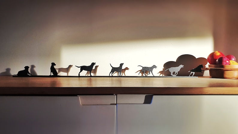 May include: A row of black metal dog silhouettes on a wooden shelf. The silhouettes are of different breeds and sizes, and some are running while others are standing. There is a cloud-shaped metal silhouette behind the dogs, and a bowl of red apples is on the shelf to the right.
