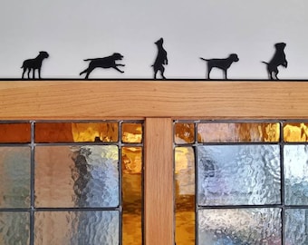 May include: A black metal decorative topper featuring six distinct dog silhouettes in various poses, including standing, running, and sitting, mounted on a thin horizontal bar designed to rest atop a wooden door frame or window ledge.
