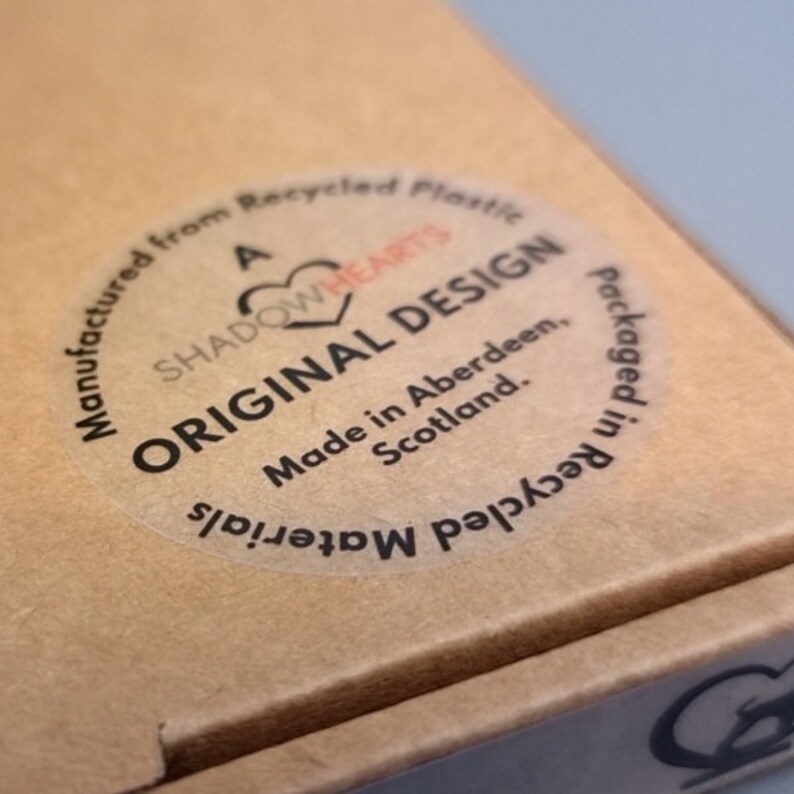 Puede incluir: Una caja de cart&oacute;n color canela con una pegatina circular semitransparente con texto negro que dice: A SHADOWHEARTS ORIGINAL DESIGN, Made in Aberdeen, Scotland, Manufactured from Recycled Plastic y Packaged in Recycled Materials.