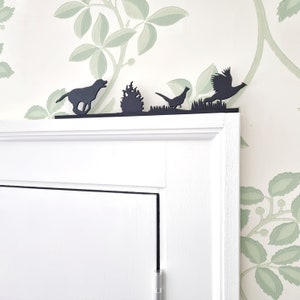 Könnte beinhalten: Schwarze Silhouette Wanddekoration mit einem Hund, einem Busch und zwei Vögeln. Die Silhouetten sind auf einem weiß lackierten Holzregal montiert.