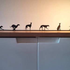 May include: A row of seven black metal silhouettes of greyhounds in various poses, including running, standing, and sitting, displayed on a wooden surface.