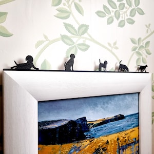 May include: A white picture frame with a black mount and a seascape painting inside. Five black silhouette dog figurines are placed on top of the frame.