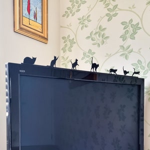 Op de afbeelding: Zwarte metalen katten-silhouetten zijn gerangschikt op een zwart tv-scherm. De silhouetten staan in een rij en lopen over de bovenkant van het scherm. De tv staat op een standaard en bevindt zich voor een muur met een bloemenpatroon.