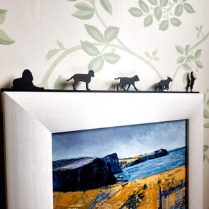 May include: A white picture frame with a black and white landscape painting of a rocky coastline.  Five black metal silhouettes of dogs are mounted on the top of the frame.