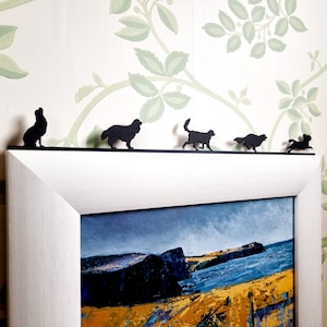 Op de afbeelding: Een wit fotolijst met een zwarte passe-partout en een landschapschilderij van een rotsachtige kustlijn met een blauwe zee. Vijf zwarte metalen silhouetten van honden zijn bovenaan de lijst bevestigd.