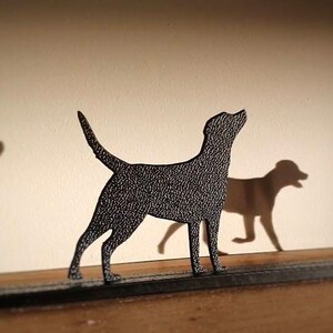 May include: A black metal silhouette of a dog standing on a wooden surface. The dog is looking up and has its tail raised. There are two other dog silhouettes in the background, one on the left and one on the right.