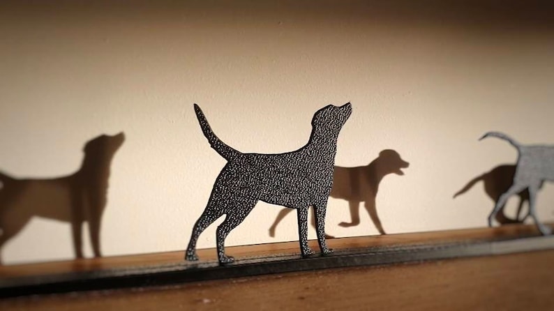 May include: A black metal silhouette of a dog standing on a wooden surface. The dog is looking up and has its tail raised. There are two other dog silhouettes in the background, one on the left and one on the right.