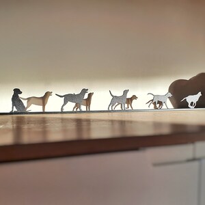 May include: A metal silhouette of a pack of dogs running in a line. The silhouettes are cut out of metal and are mounted on a wooden shelf. The silhouettes are all facing the same direction and are in a line.