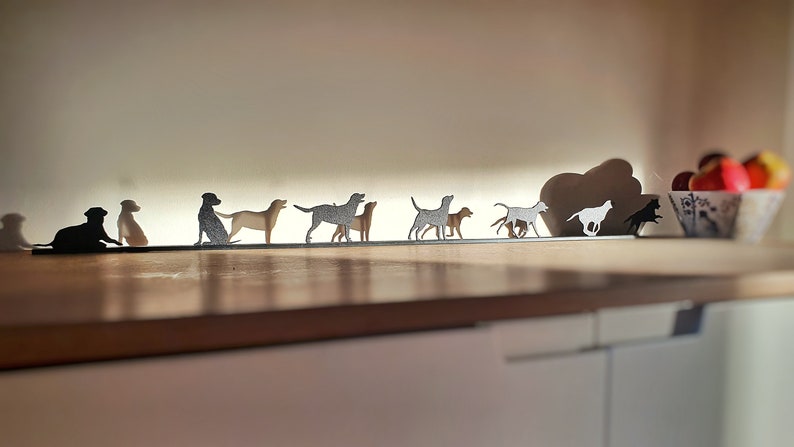 May include: A metal silhouette of a pack of dogs running in a line. The silhouettes are cut out of metal and are mounted on a wooden shelf. The silhouettes are all facing the same direction and are in a line.