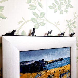Può includere: Una cornice bianca con un passepartout nero e un dipinto di paesaggio in bianco e nero all'interno. Ci sono cinque sagome di cani in metallo nero sulla parte superiore della cornice.