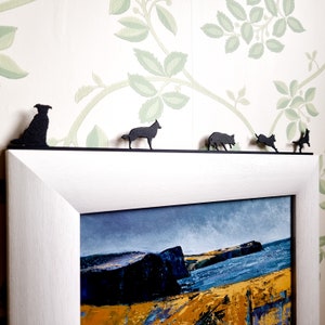 Könnte beinhalten: Ein weißer Bilderrahmen mit schwarzem Passepartout und einem Landschaftsbild einer felsigen Küste mit blauem Wasser. Fünf schwarze Metall-Silhouetten von Hunden sind oben auf dem Rahmen platziert.